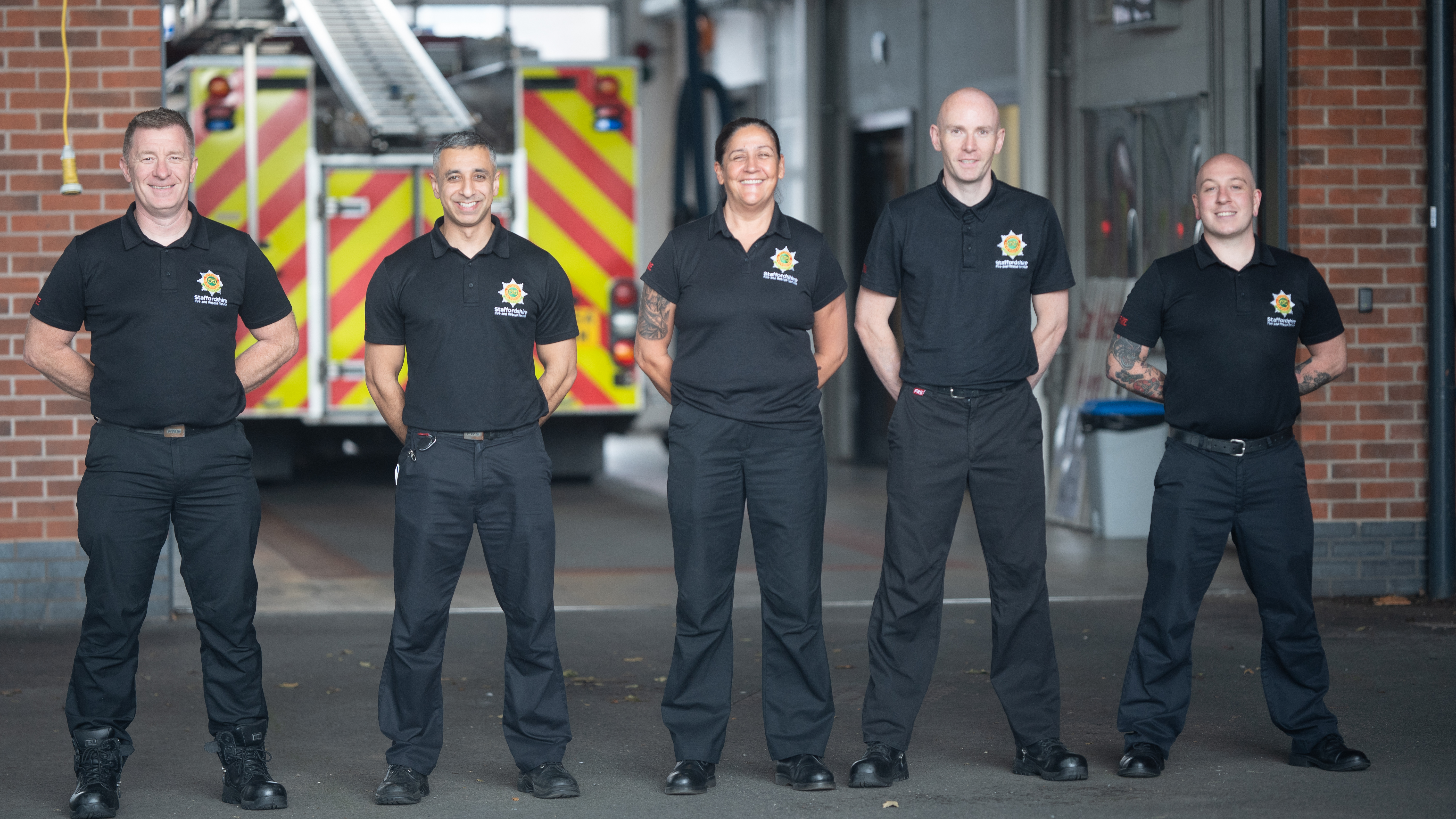 Five firefighters stood in a line in front of a fire appliance bay facing the camera smiling